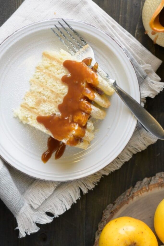 Slice of cake with caramel sauce on a white plate, set on a rustic table with a fork and apples.