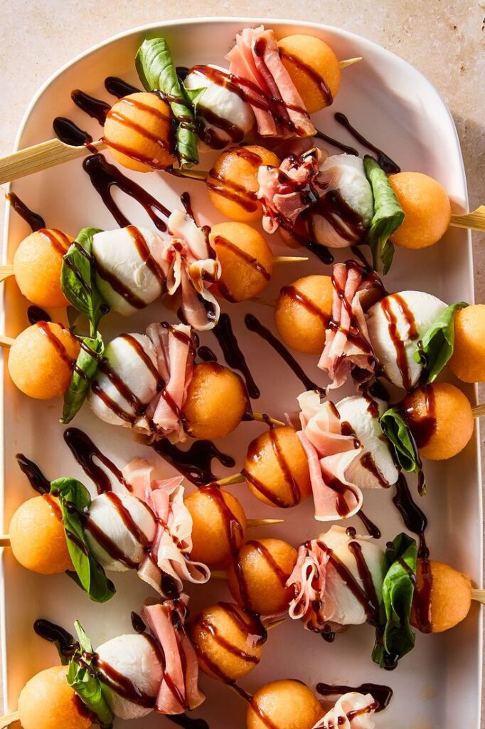 Melon, mozzarella, prosciutto, and basil skewers drizzled with balsamic glaze on a white plate.