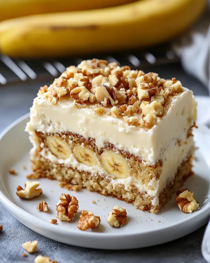 Creamy banana walnut cake slice on white plate, with fresh bananas in background. Perfect dessert for any occasion.