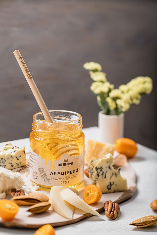 Jar of acacia honey with cheese, nuts, and fruits on a wooden board, accented by flowers in the background.