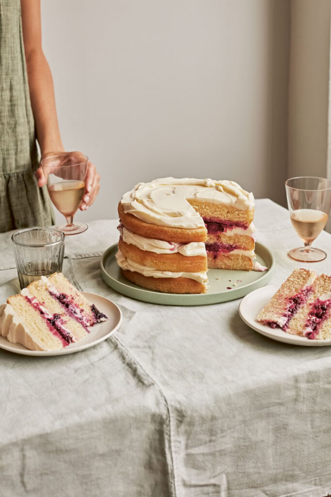 Layered cake with berry filling and icing on a table, accompanied by two slices on plates and glasses of white wine.