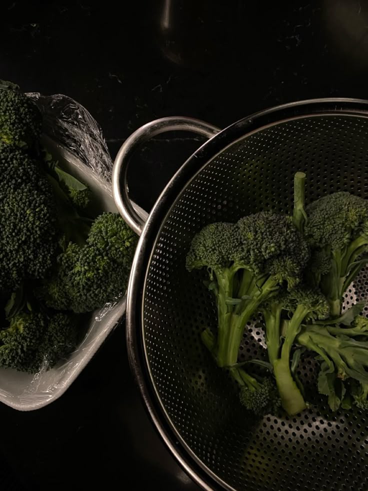 Fresh broccoli in a metal colander, ready for cooking on a dark kitchen countertop.