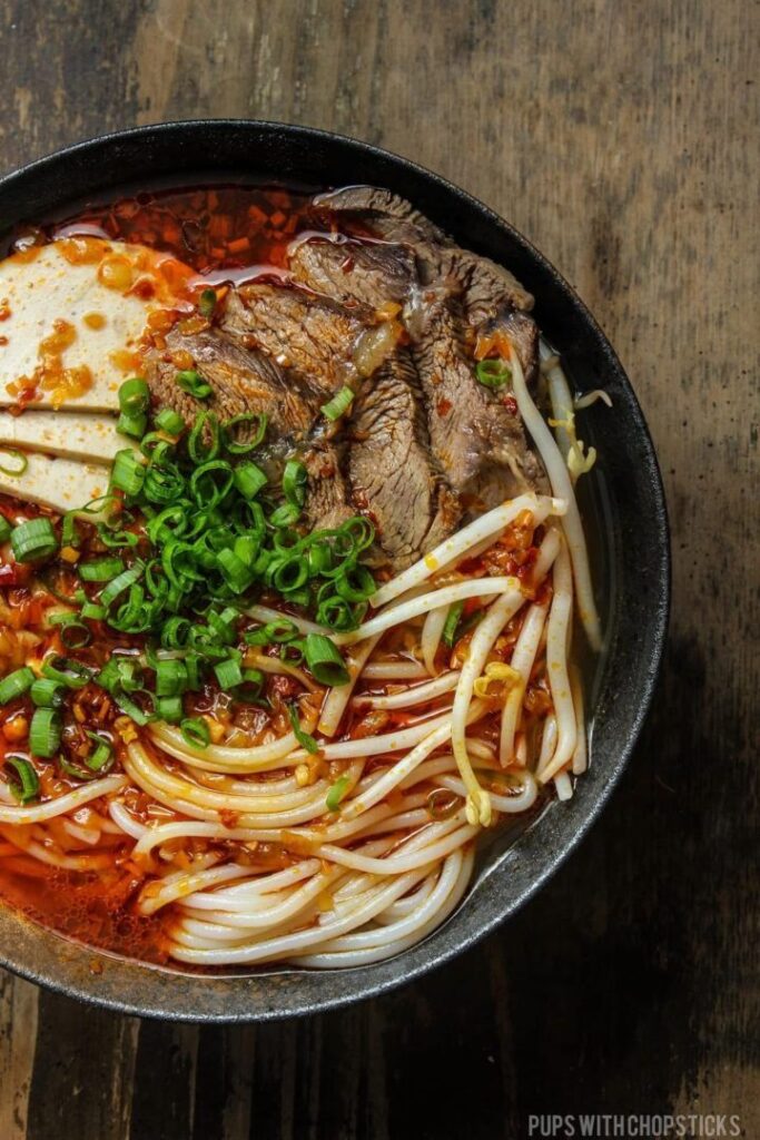 Spicy beef noodle soup with tofu, bean sprouts, and green onions in a black bowl on a wooden table.
