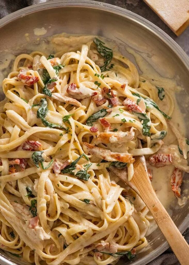 Creamy fettuccine pasta with chicken, spinach, and sun-dried tomatoes in a skillet with wooden spoon.