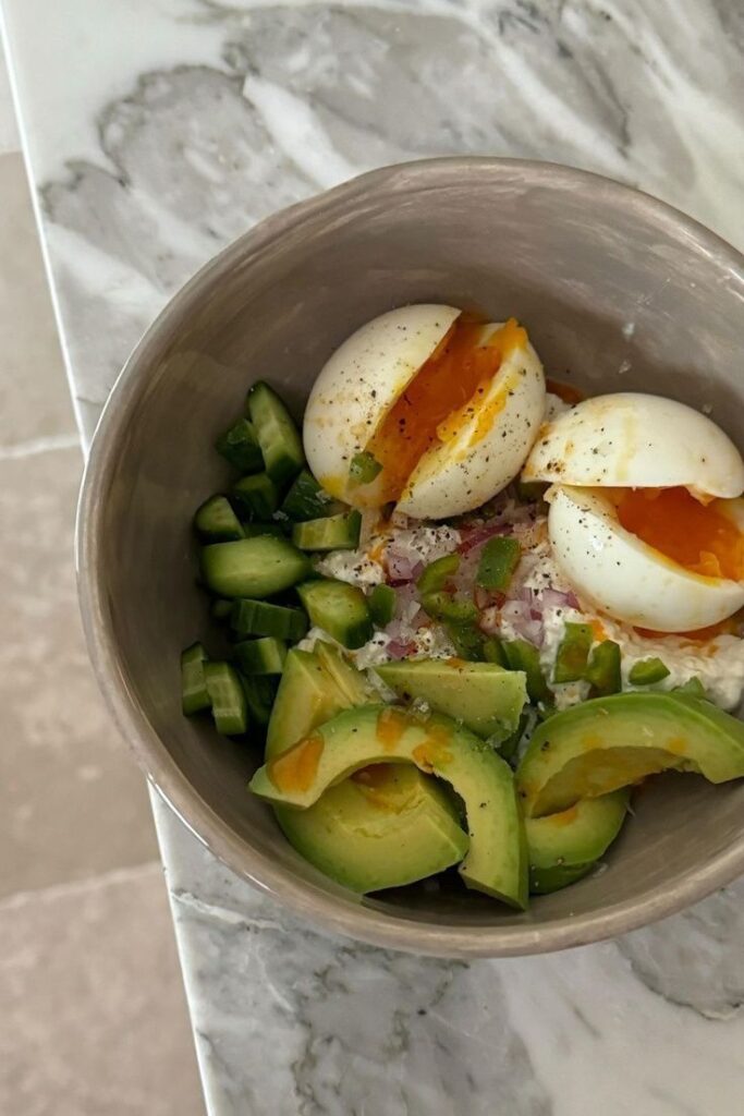 Fresh breakfast bowl with avocado, soft-boiled eggs, cucumbers, and peppers on a marble table. Healthy meal.