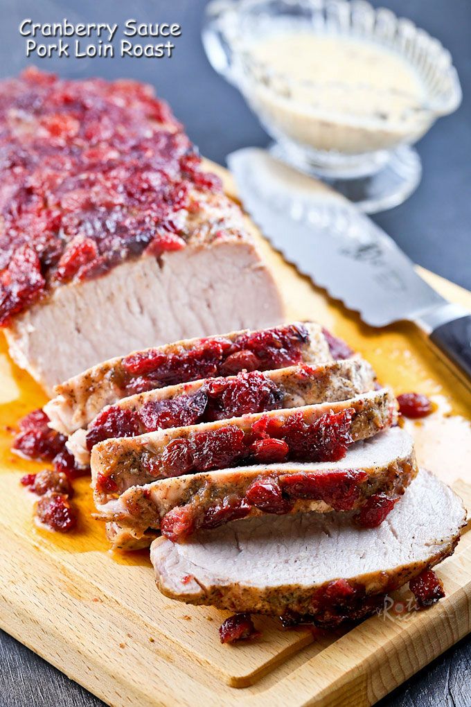 Sliced pork loin roast with cranberry sauce glaze on a wooden board, served with creamy sauce and knife.