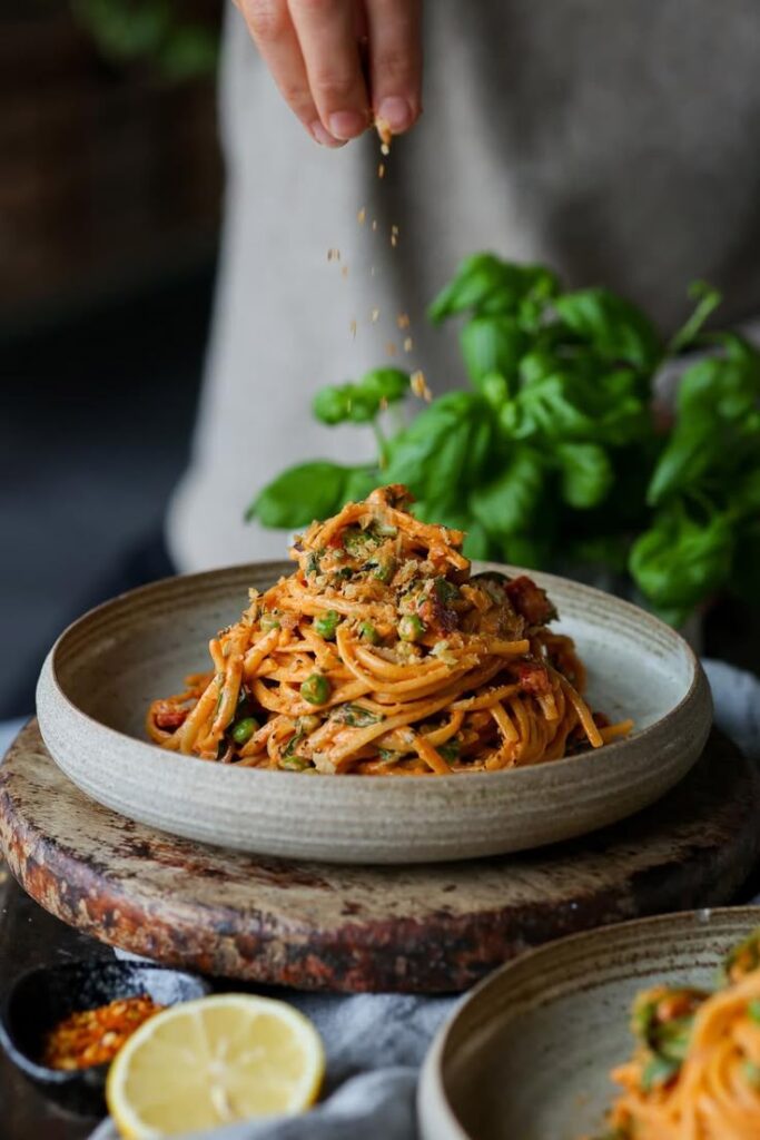 Hand sprinkling seasoning on a plate of creamy pasta with vegetables, garnished with fresh herbs and lemon slice.