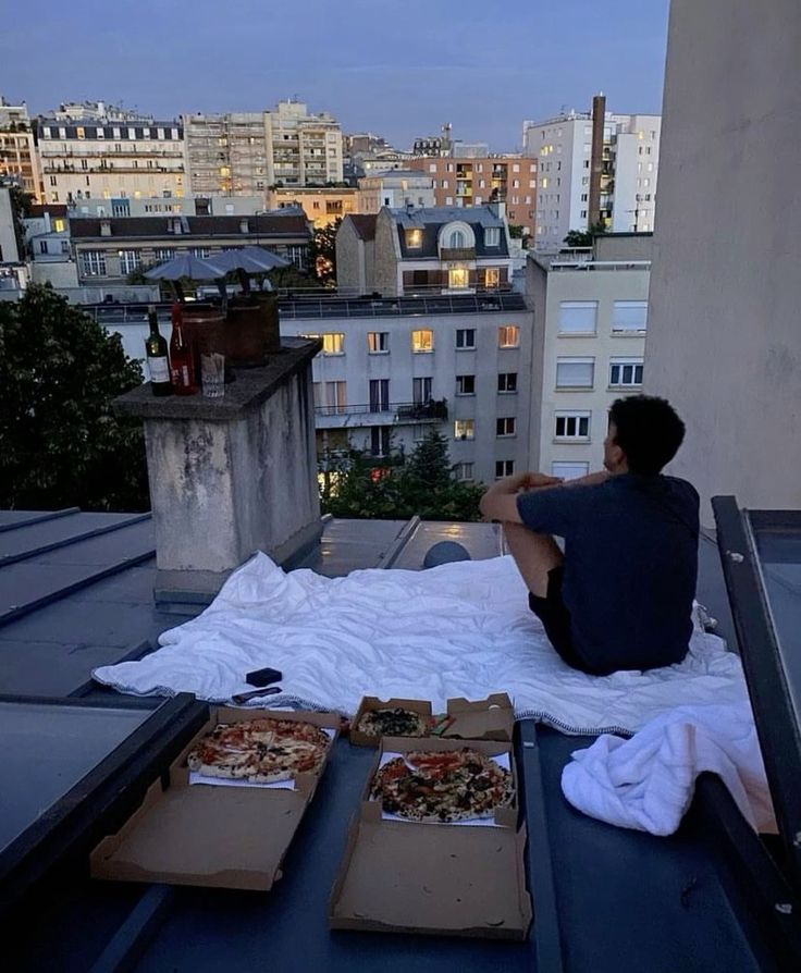 Person enjoying rooftop picnic with pizzas at sunset in a cityscape setting.