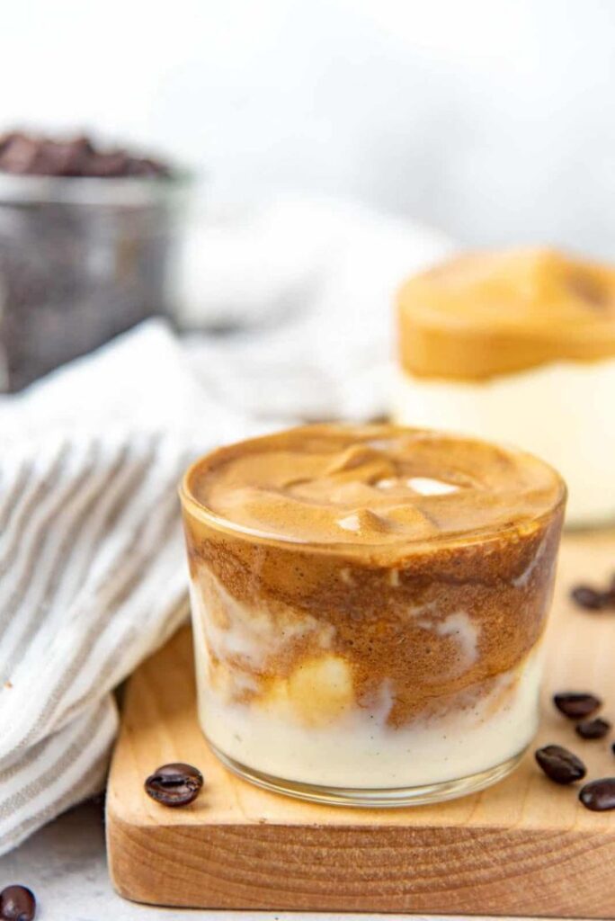 Creamy dalgona coffee on a wooden board with scattered coffee beans, perfect for a trendy homemade caffeine fix.