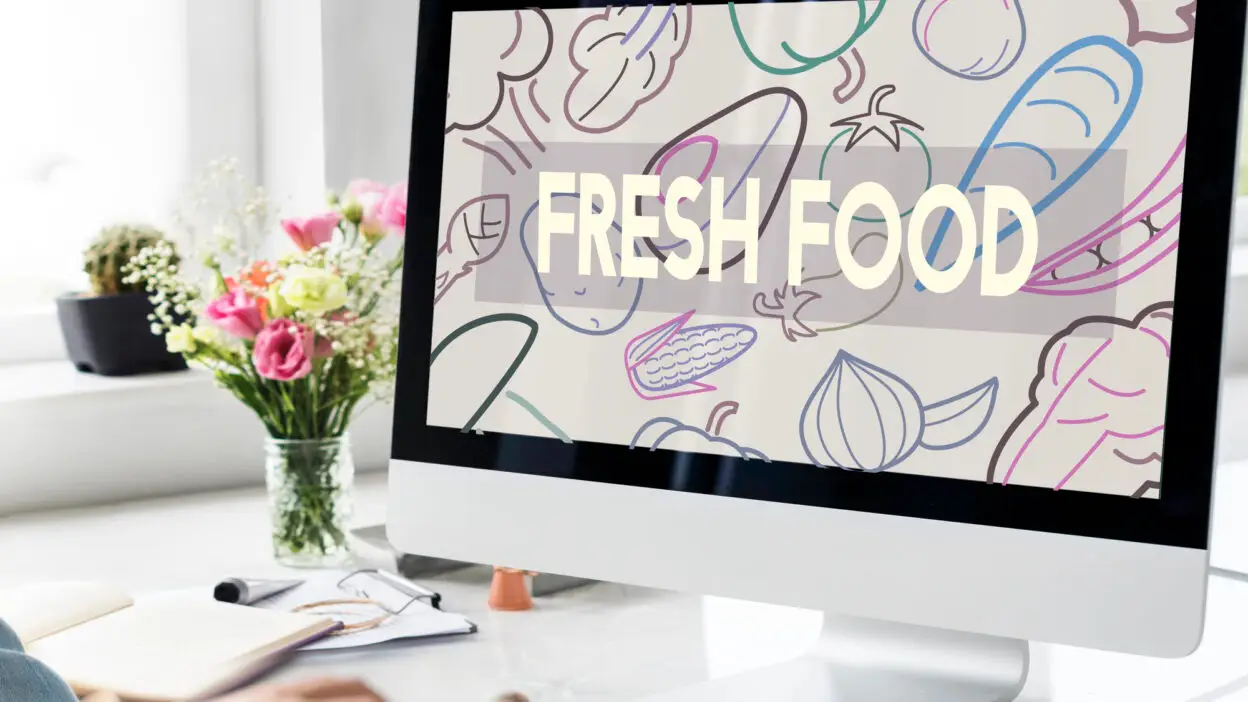Computer screen showing Fresh Food with vegetable illustrations, set on a desk with flowers and a cup of tea.
