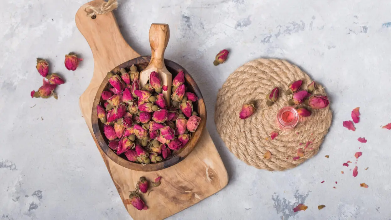 Wooden bowl of dried pink rosebuds on a cutting board with decorative mat and glass bottle, on a textured background.