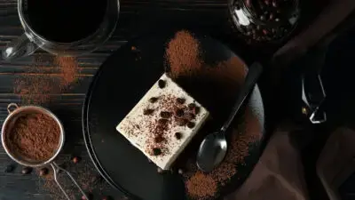 Tiramisu dessert with coffee beans on a black plate, surrounded by cocoa powder and a cup of coffee.