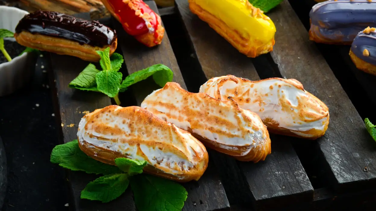 Assorted colorful eclairs with mint leaves on a wooden platter, featuring chocolate, red, yellow, and white toppings.
