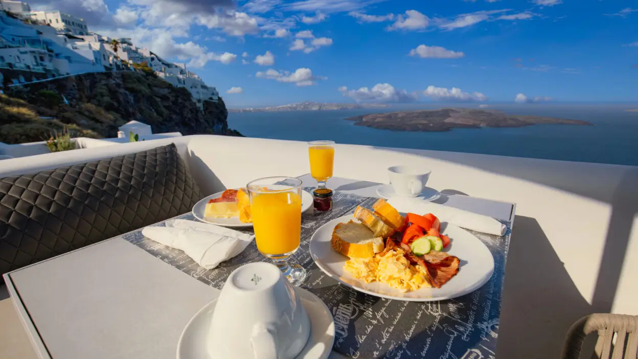 Breakfast with orange juice overlooking Santorini cliffs and sea under a blue sky with clouds.