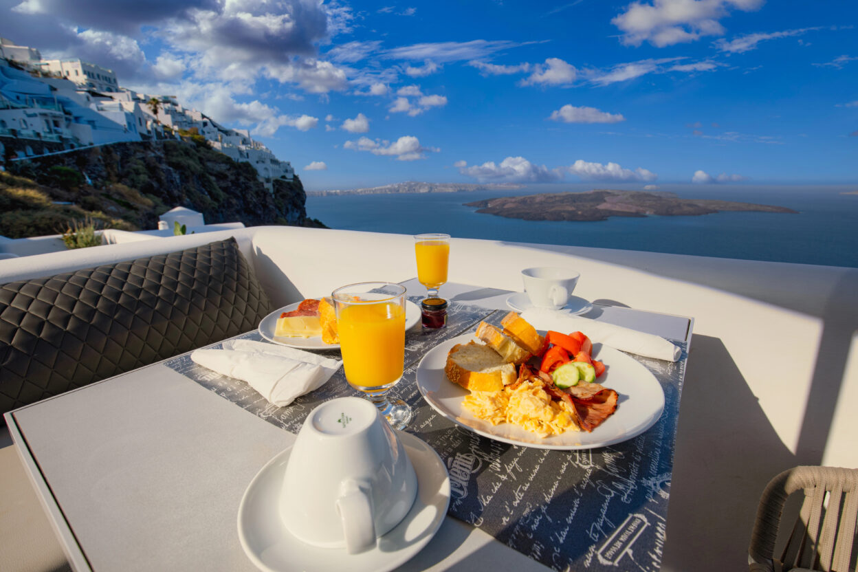 Breakfast with orange juice overlooking Santorini cliffs and sea under a blue sky with clouds.