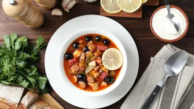 Hearty stew with sausage, olives, and lemon slice in a white bowl, surrounded by bread and fresh herbs on a wooden table.