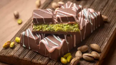 Chocolate bars with pistachio filling, decorated with drizzled frosting, on a wooden board with scattered pistachios.