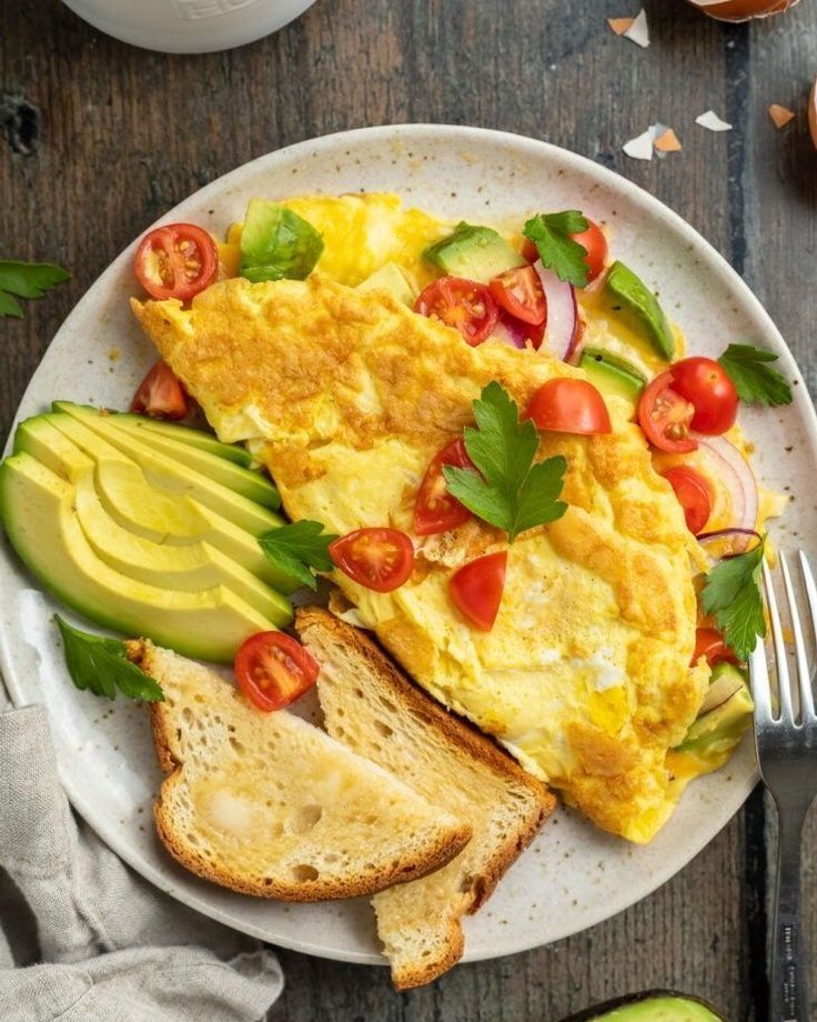 Delicious omelette with avocado, tomatoes, toast, and herbs on a white plate. Perfect healthy breakfast option.