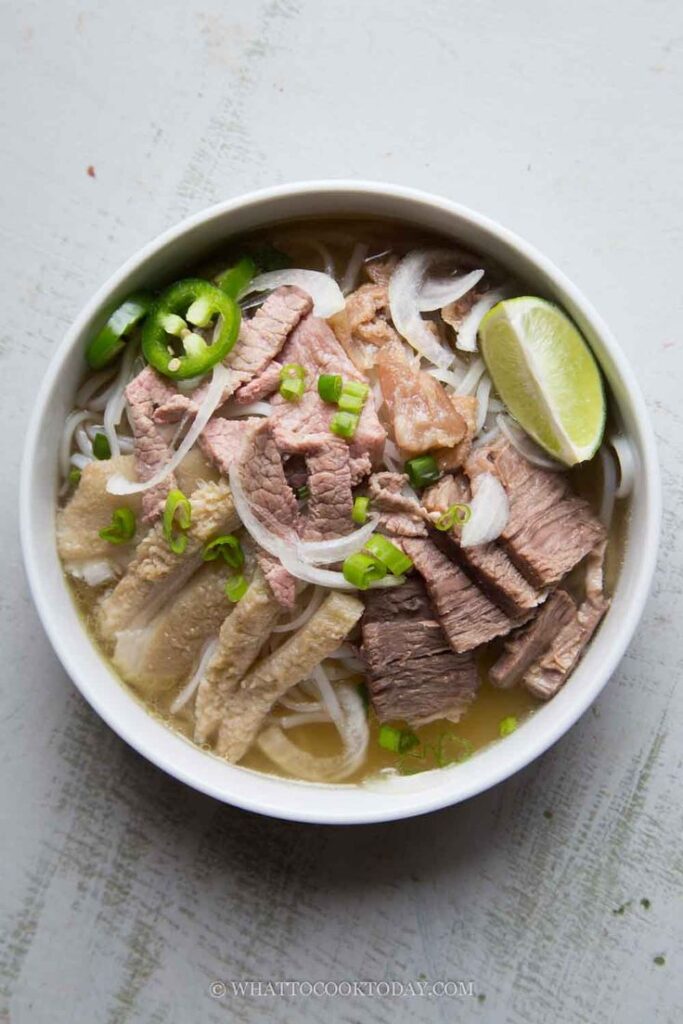 Bowl of Vietnamese pho with beef, fresh lime, jalapeño, onion, and herbs on a light background.