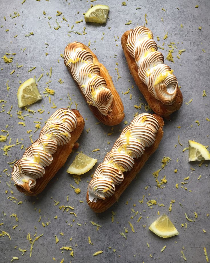 Lemon meringue eclairs topped with swirled frosting, surrounded by fresh lemon slices and zest on a grey background.