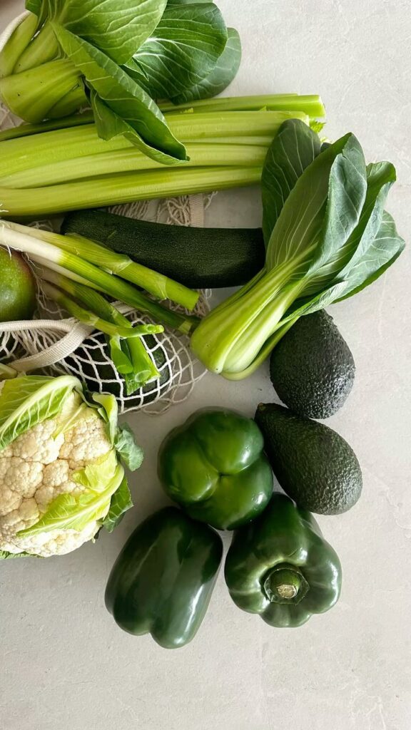 Fresh green vegetables including celery, bok choy, avocado, bell peppers, and cauliflower on a light surface.