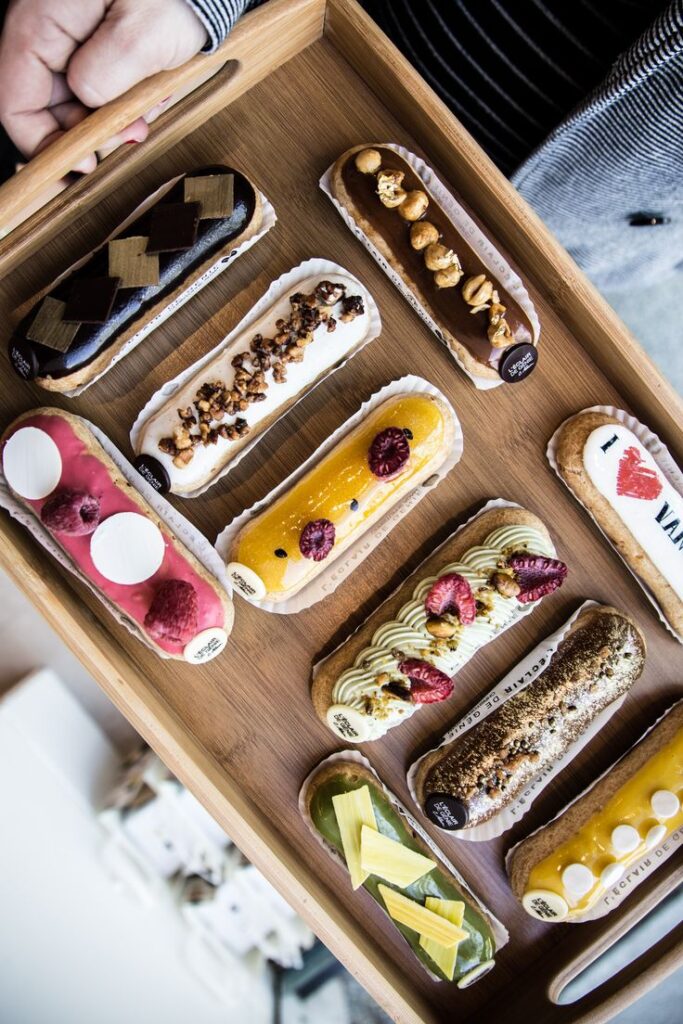 A tray of assorted colorful eclairs with various toppings, held by a hand, on a wooden surface.