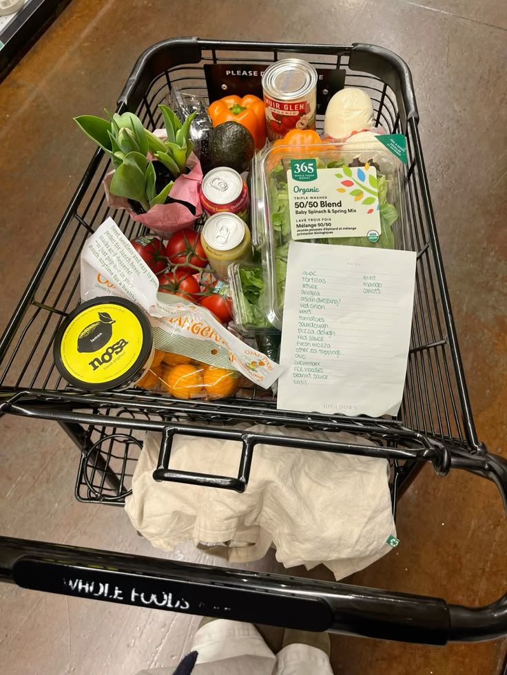 Shopping cart filled with fresh produce, canned goods, and a grocery list at a supermarket.