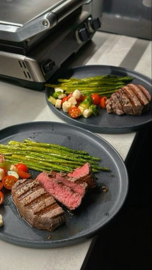 Grilled steak with asparagus and vegetable medley on black plates next to a stainless steel grill.