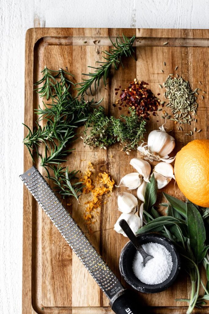 Fresh herbs, garlic, orange zest, and spices on a wooden board with a grater and salt bowl, perfect for cooking.