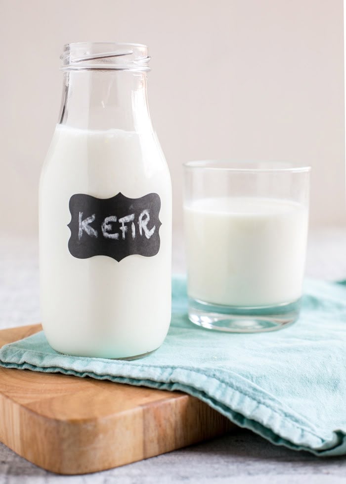 Glass bottle of kefir with a chalk label and a glass of kefir on a wooden board with a teal cloth.