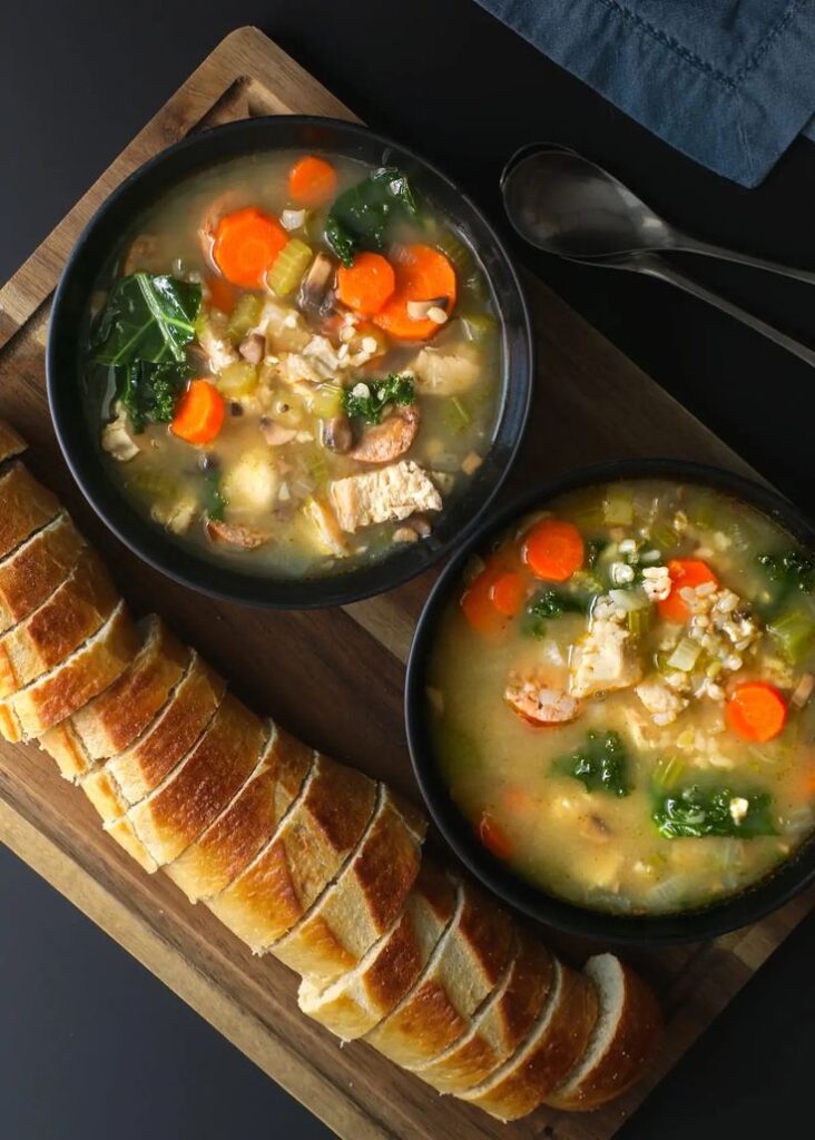 Bowl of hearty chicken vegetable soup with sliced baguette on a wooden board, perfect for a cozy meal.