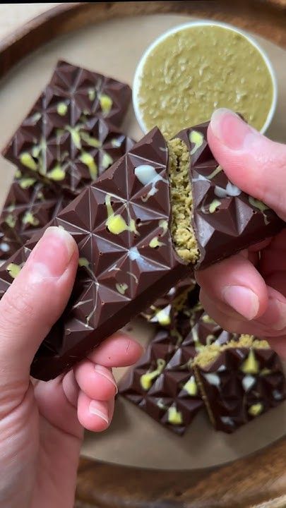 Person breaking a geometric chocolate bar with green filling, next to a bowl of green sauce on a wooden tray.