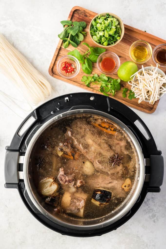 Instant Pot pho broth cooking with fresh herbs and spices on a wooden board, alongside rice noodles and toppings.
