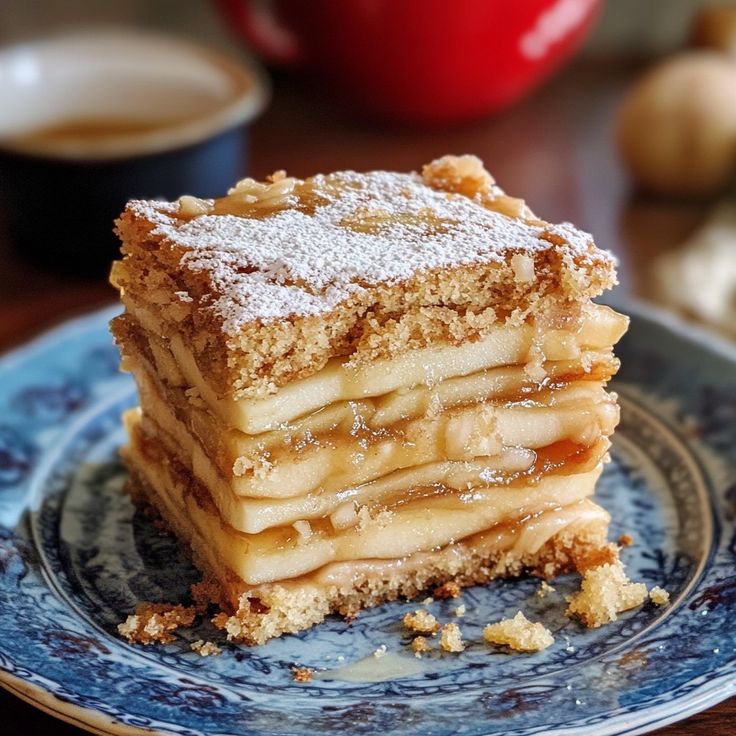 Layered apple cake slice on a blue floral plate, topped with powdered sugar, with coffee in the background.