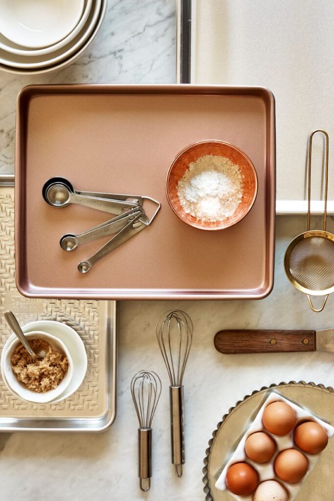 Baking essentials arranged on a counter: tray, measuring spoons, brown sugar, eggs, whisks, and sifter.
