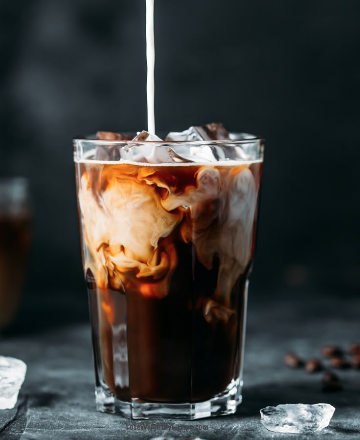 Iced coffee with milk swirling in a glass, set against a dark background with ice cubes nearby.