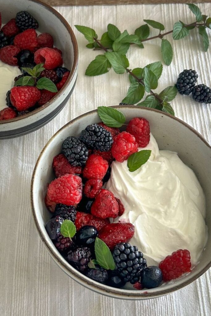 Bowl of fresh raspberries, blackberries, and creamy yogurt garnished with mint leaves, on a white cloth background.