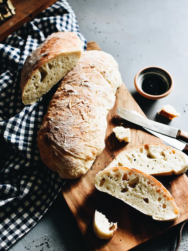 Freshly baked ciabatta bread on a wooden cutting board with a knife and olive oil dip.