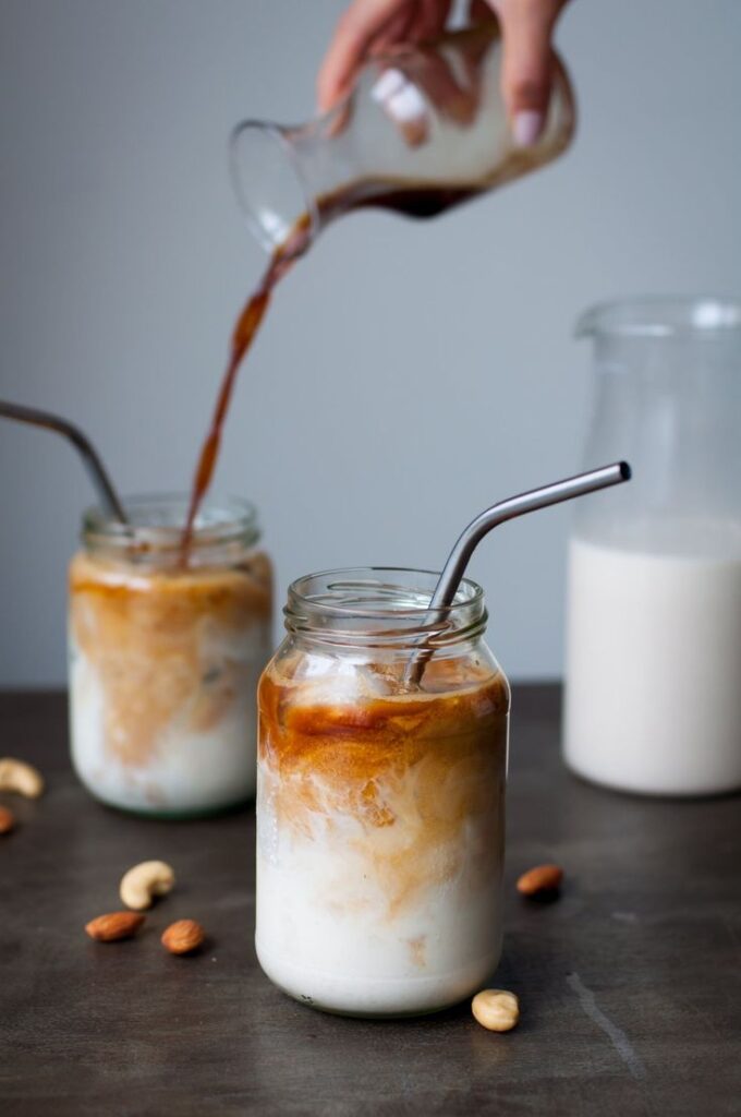Pouring coffee into iced milk jars with metal straws, surrounded by almonds and a milk jug.