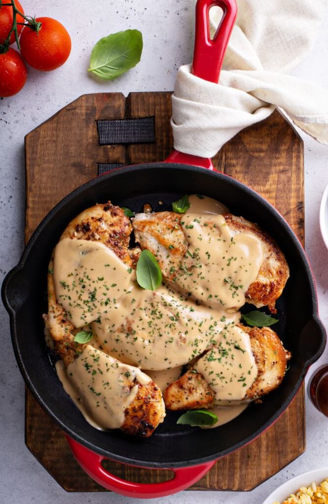 Creamy chicken breasts garnished with herbs in a red skillet on a wooden board with fresh tomatoes and basil.