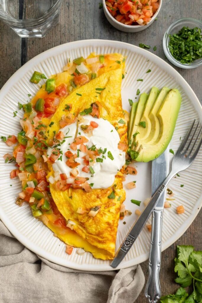 Delicious omelette topped with sour cream, salsa, and sliced avocado on a plate with cutlery.
