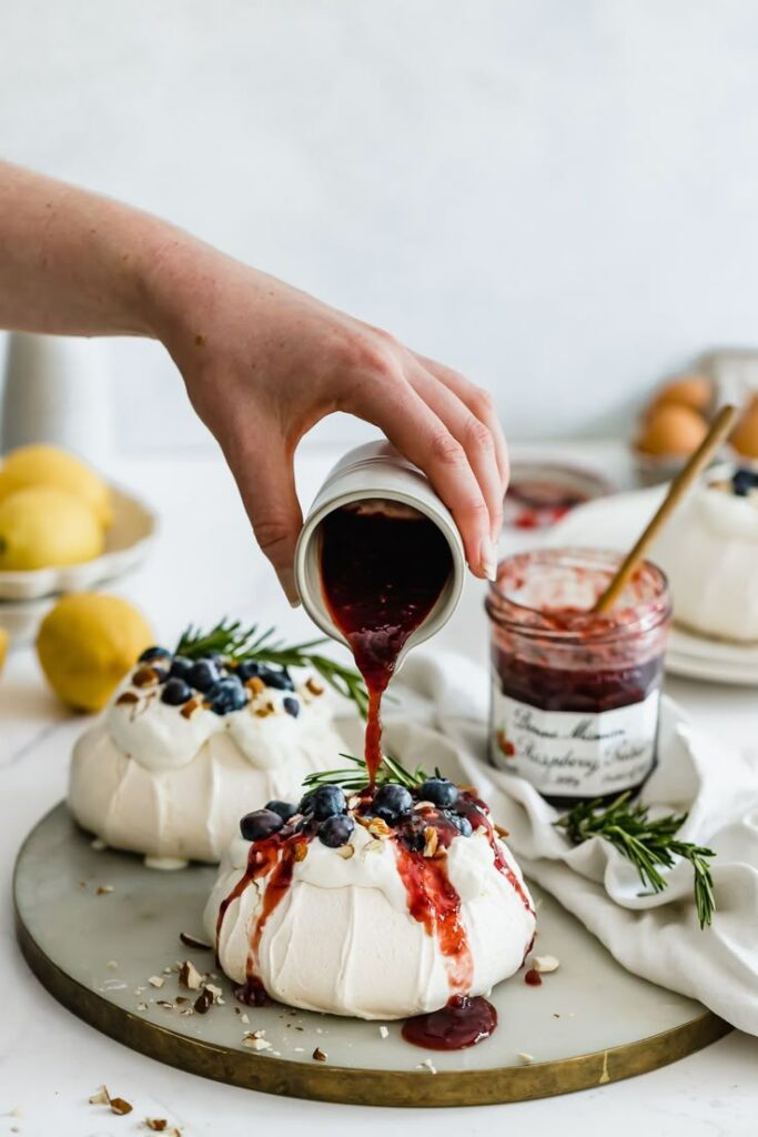Hand pouring berry sauce over pavlova topped with blueberries and nuts on a platter.