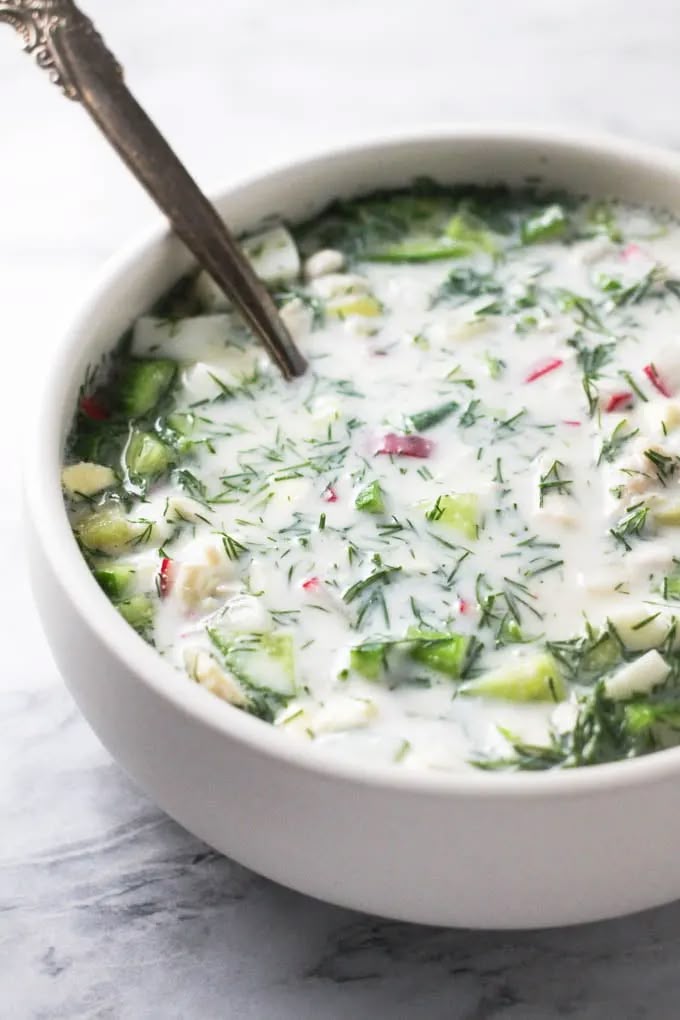 Chilled bowl of okroshka soup with fresh herbs and veggies, featuring a creamy base and a spoon, on a marble surface.