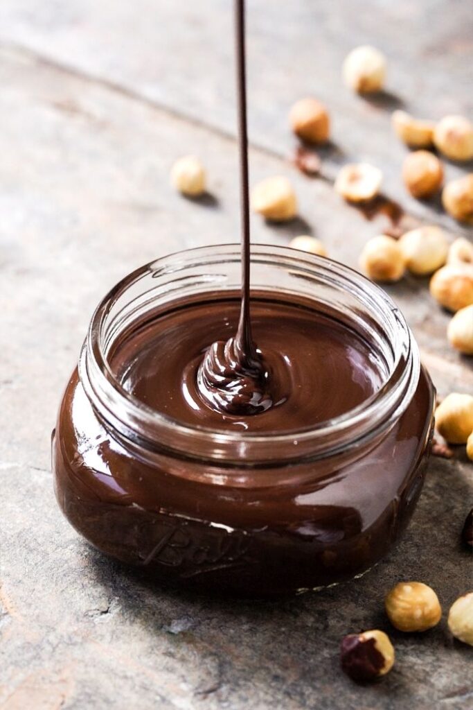 Creamy chocolate hazelnut spread being poured into a jar, surrounded by scattered hazelnuts.