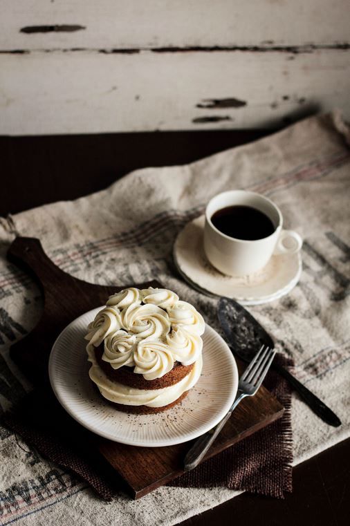 Cake topped with creamy roses served on a wooden board, next to a cup of coffee on a rustic tablecloth.