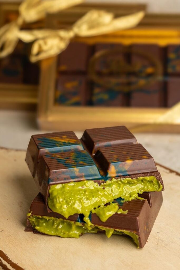 Artisan chocolate bar with vibrant green filling, painted with colorful abstract designs, resting on a wooden surface.