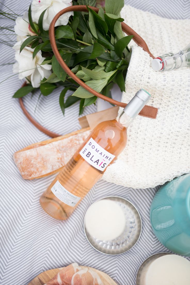Picnic setup with a bottle of rosé wine, a baguette, flowers, and a woven bag on a striped cloth.