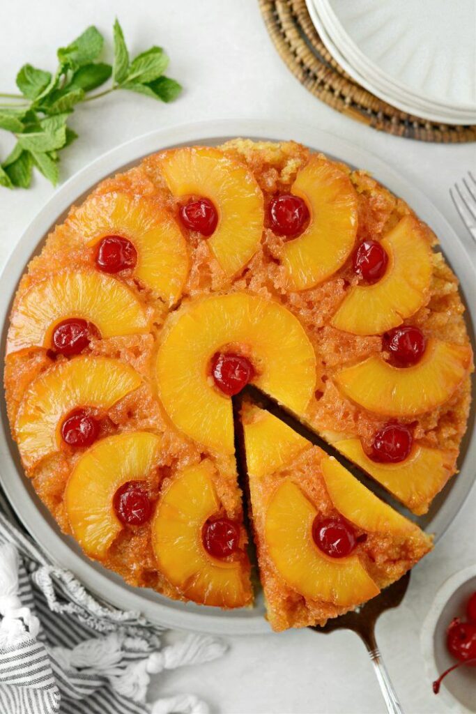 Pineapple upside-down cake with cherries on a plate, slice being served. Perfect dessert with vibrant colors.
