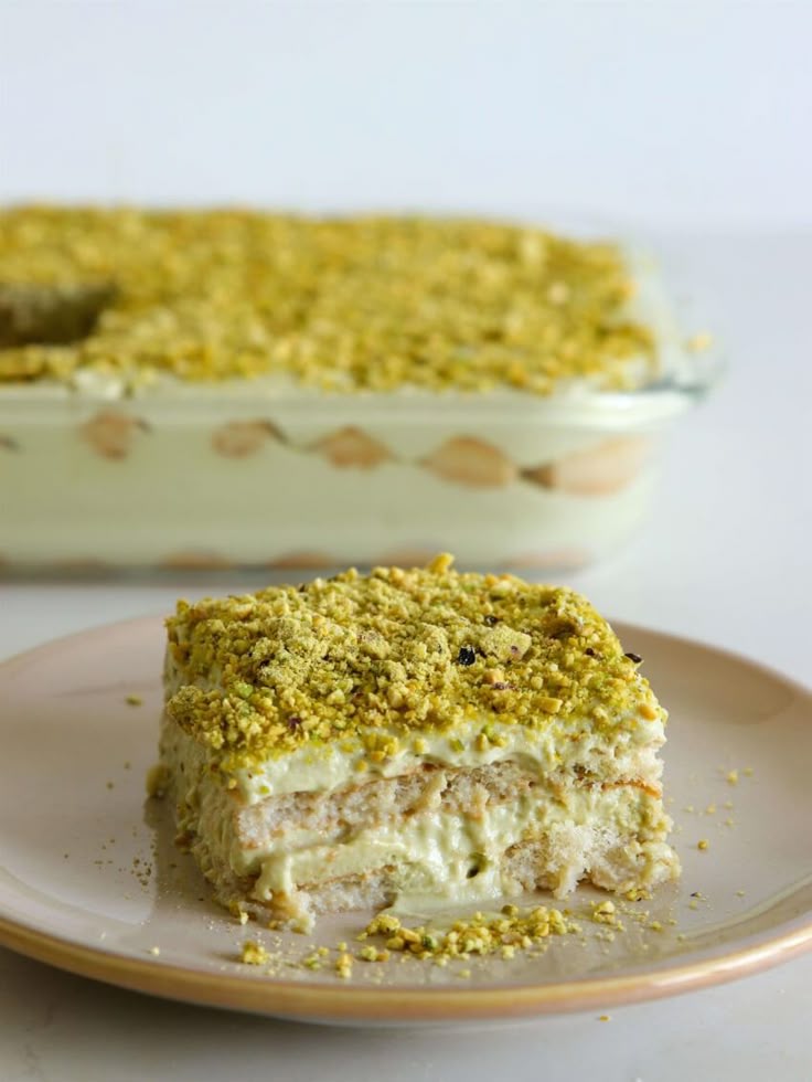 Slice of creamy pistachio tiramisu on a plate, topped with crushed pistachios, with the rest in the background.