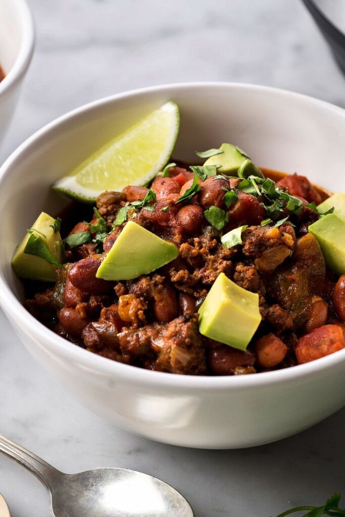 Hearty chili bowl topped with avocado, lime wedge, and fresh herbs in a white bowl on a marble surface.